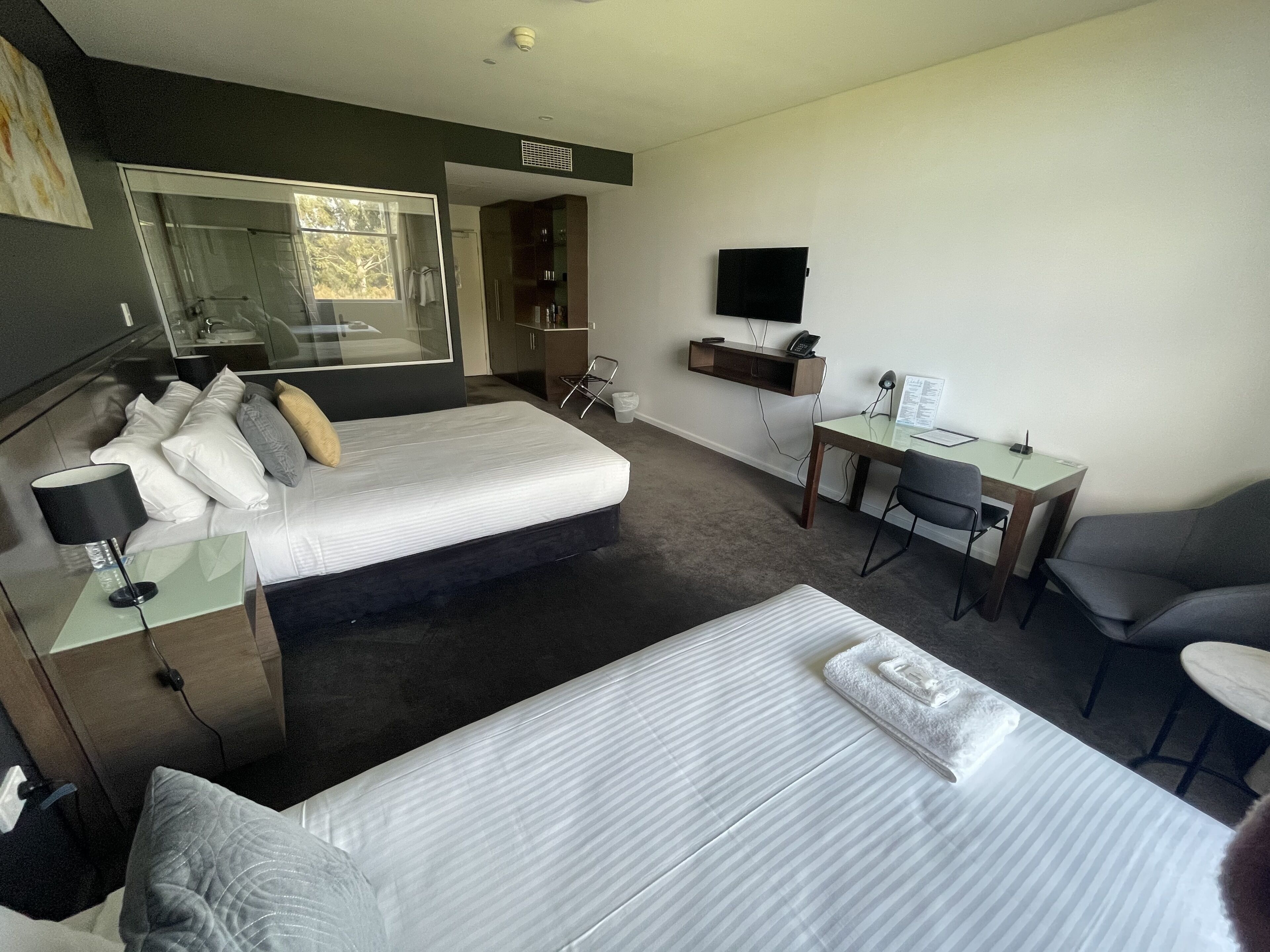 A hotel room with a bed, a desk, a chair, a TV, and a bathroom visible through a glass door.