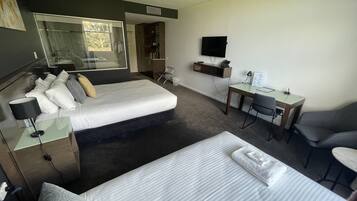 A hotel room with a bed, a desk, a chair, a TV, and a bathroom visible through a glass door.