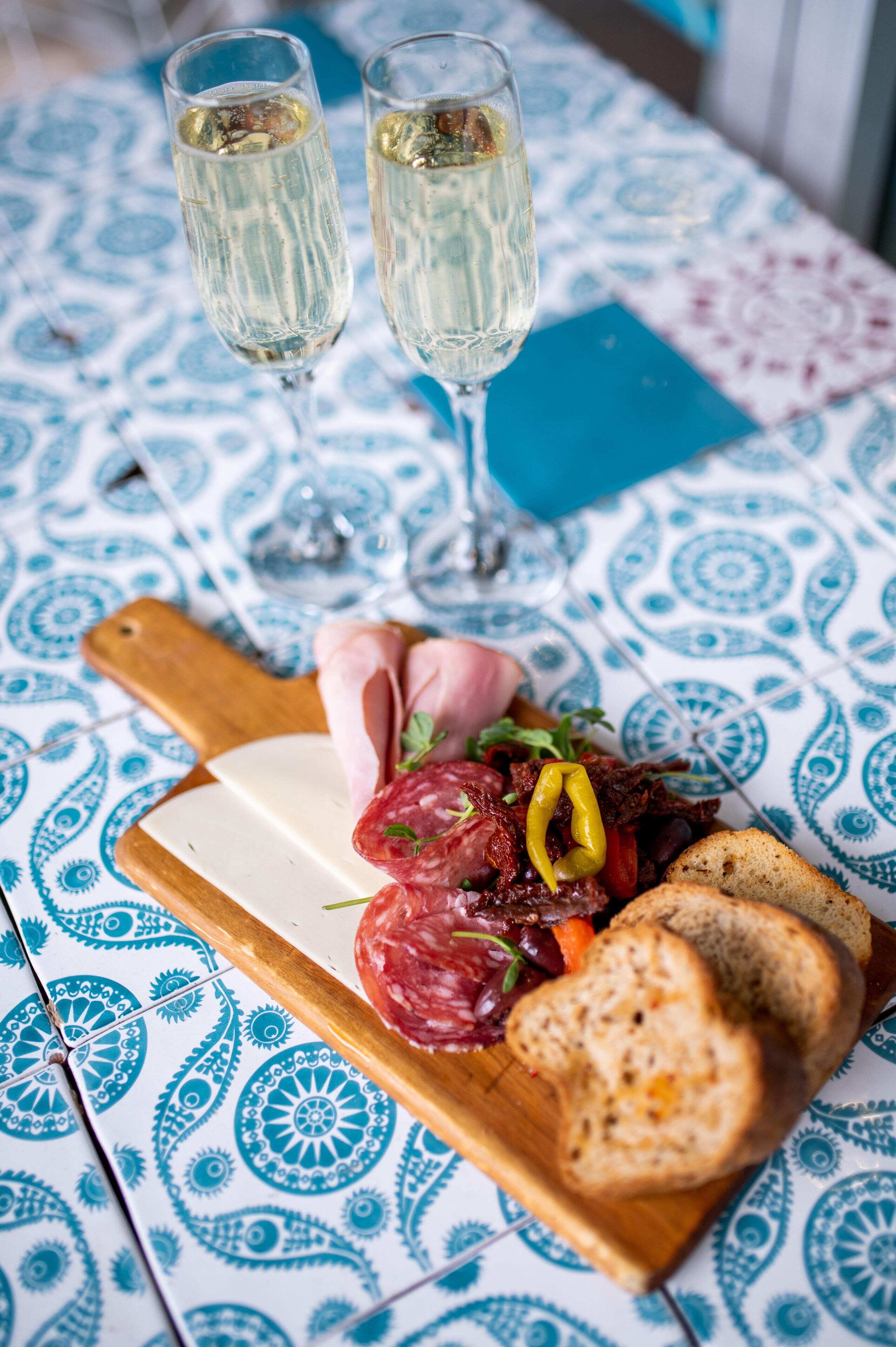 Un tavolo con una tovaglia decorata, un tagliere di legno con formaggio, salumi e pane, e due bicchieri di un liquido trasparente.