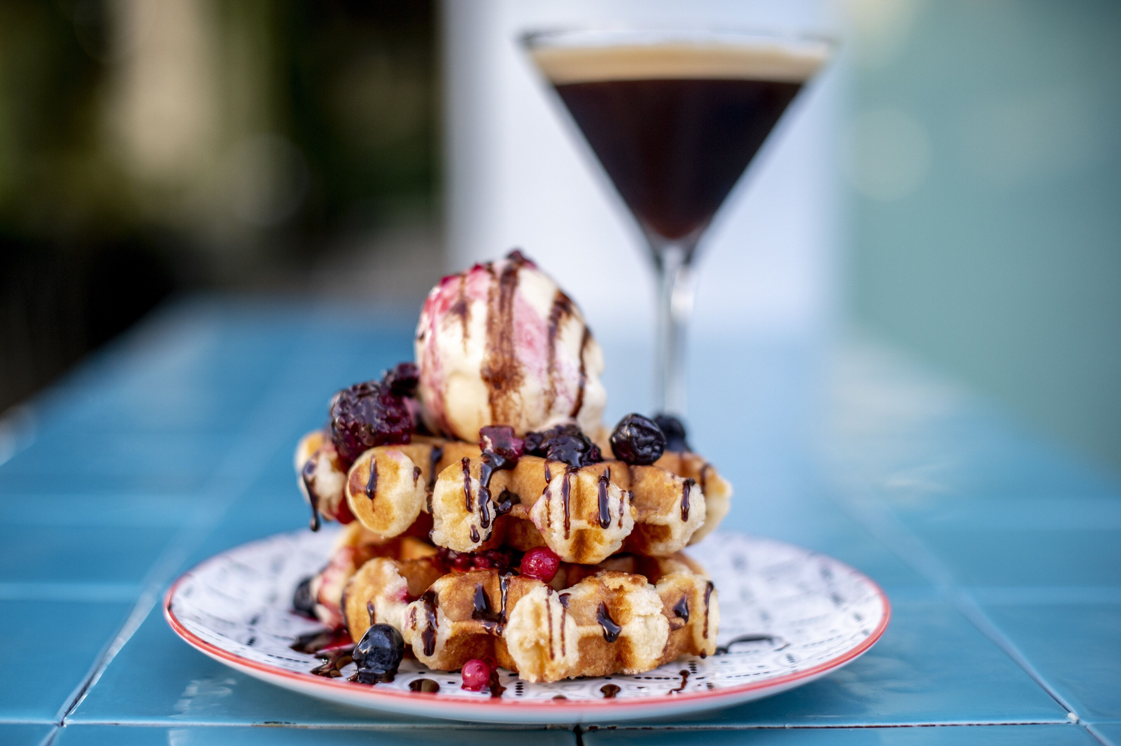 A stack of waffles with berries and syrup, a martini glass, and a cup of coffee on a blue table.