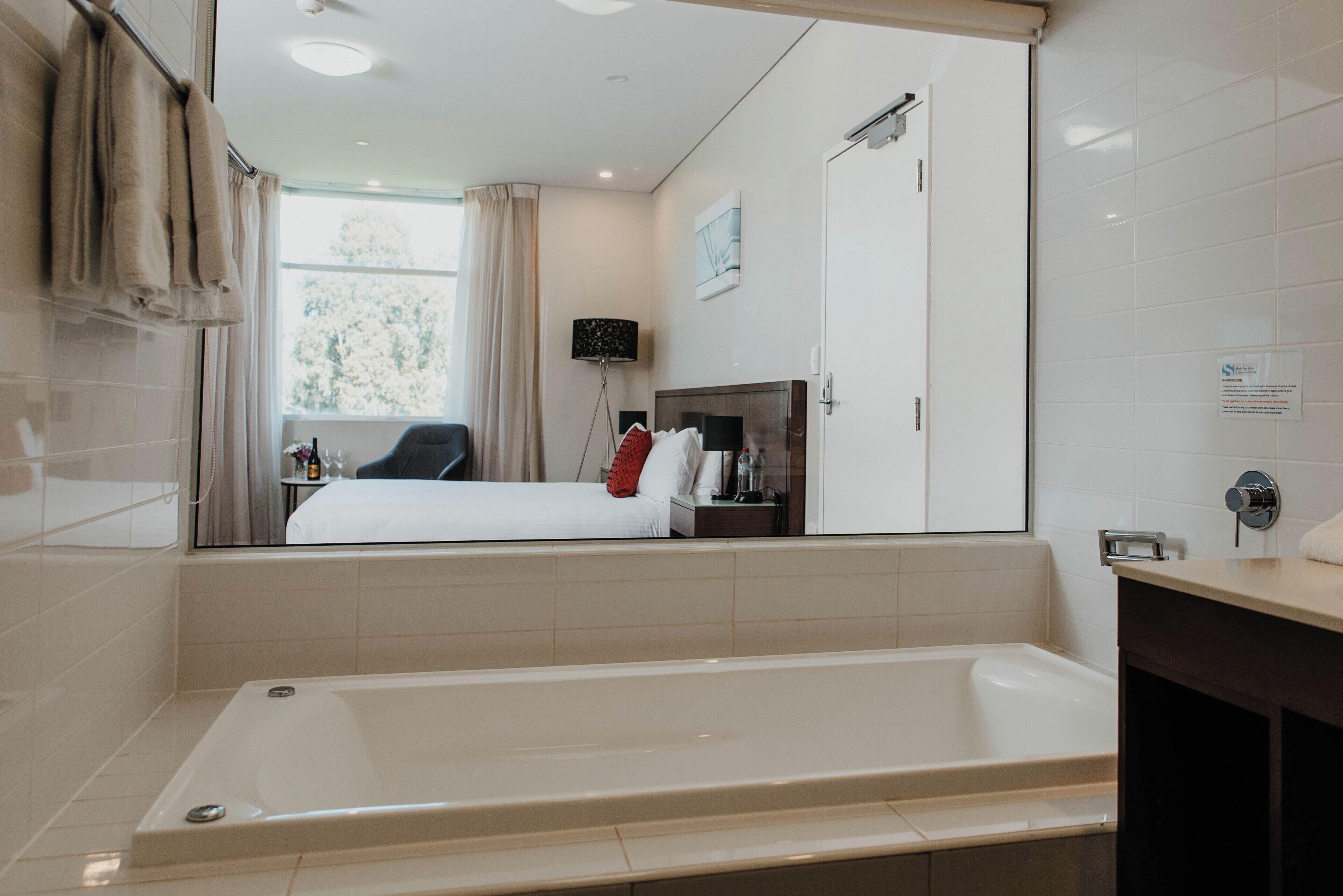 A modern bathroom with a large bathtub, a bed, and a mirror reflecting the room.