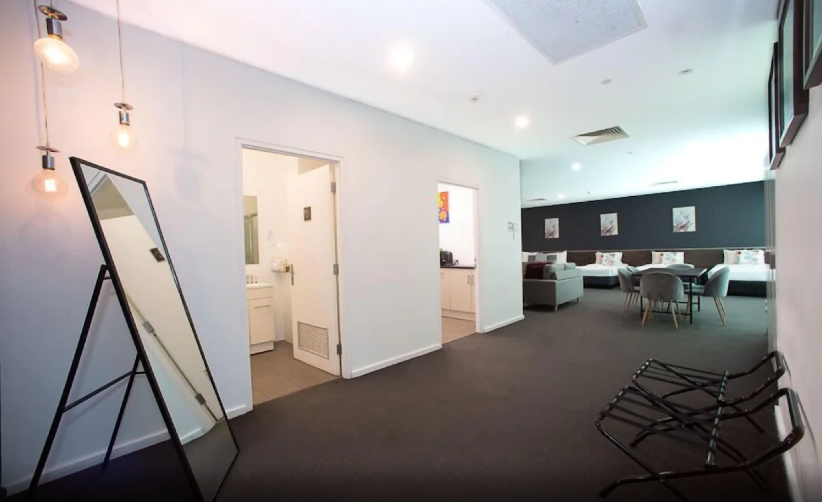 A modern hotel room with a large mirror, a dining area with a table and chairs, and a separate bathroom visible through an open door.