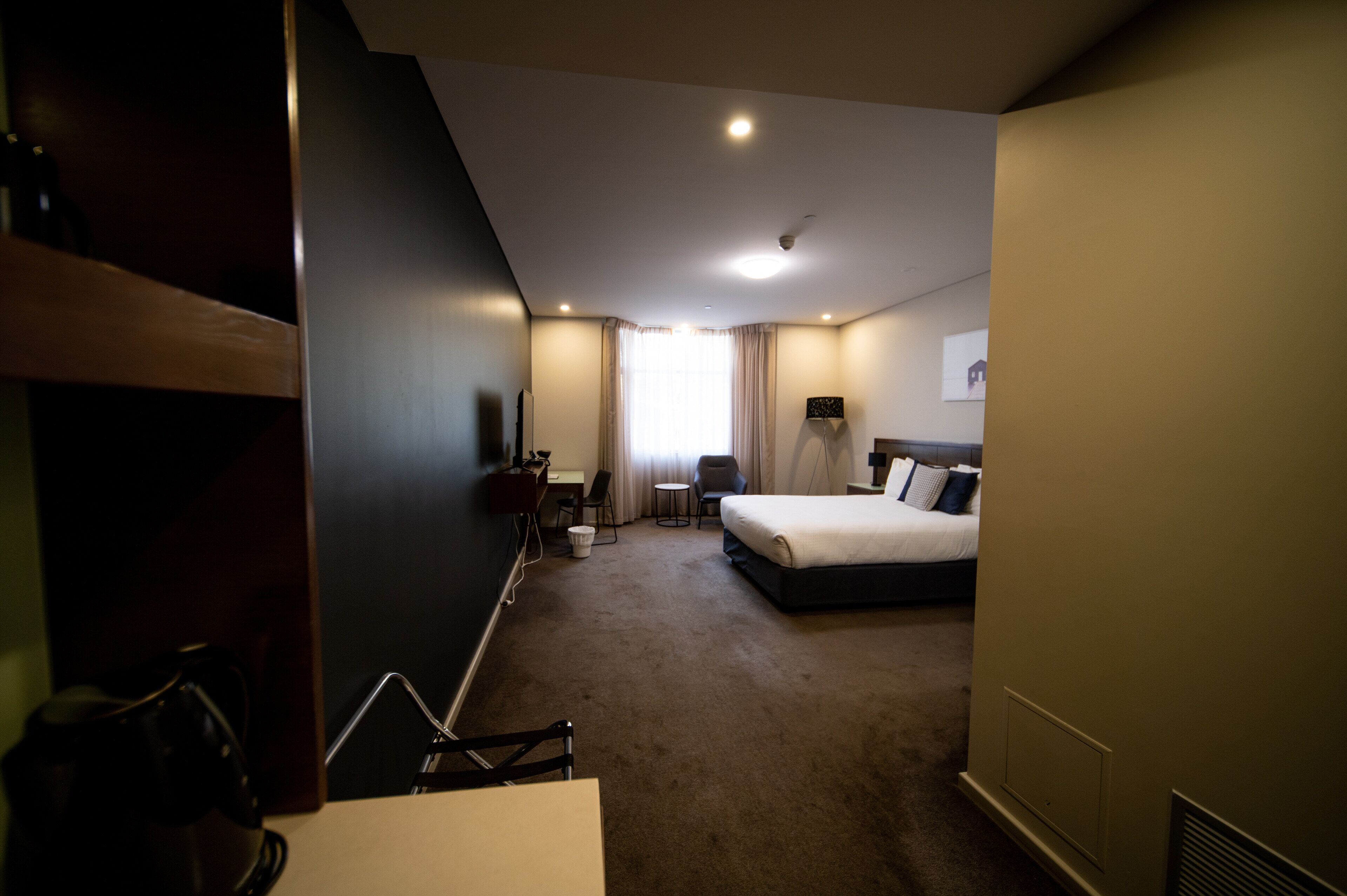Una habitación de hotel con una cama, un escritorio con silla, una mesita y un ventanal con cortinas.
