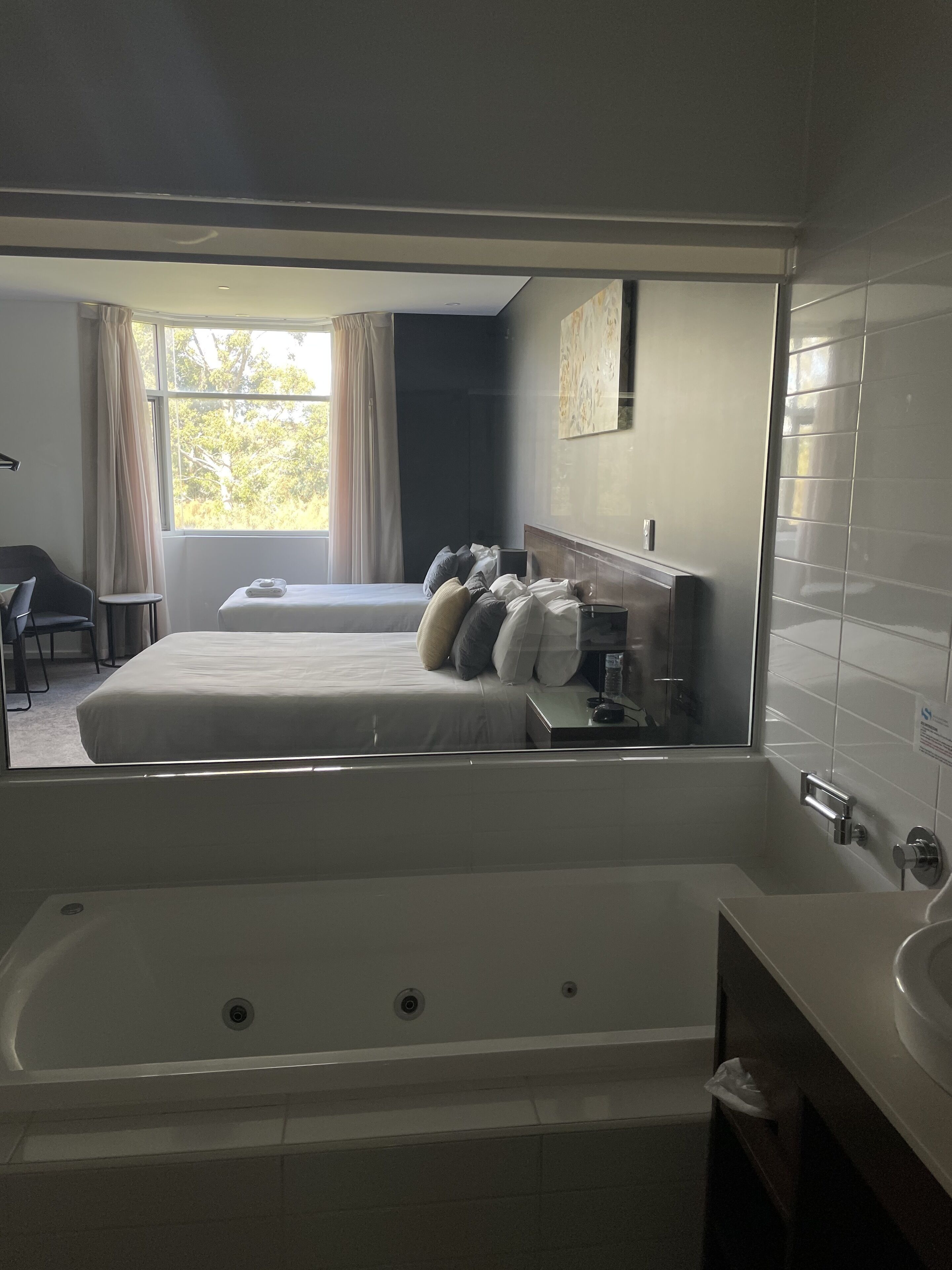 A modern hotel room with a large bed, a separate smaller bed, a bathtub, and a separate bathroom with a sink and mirror.