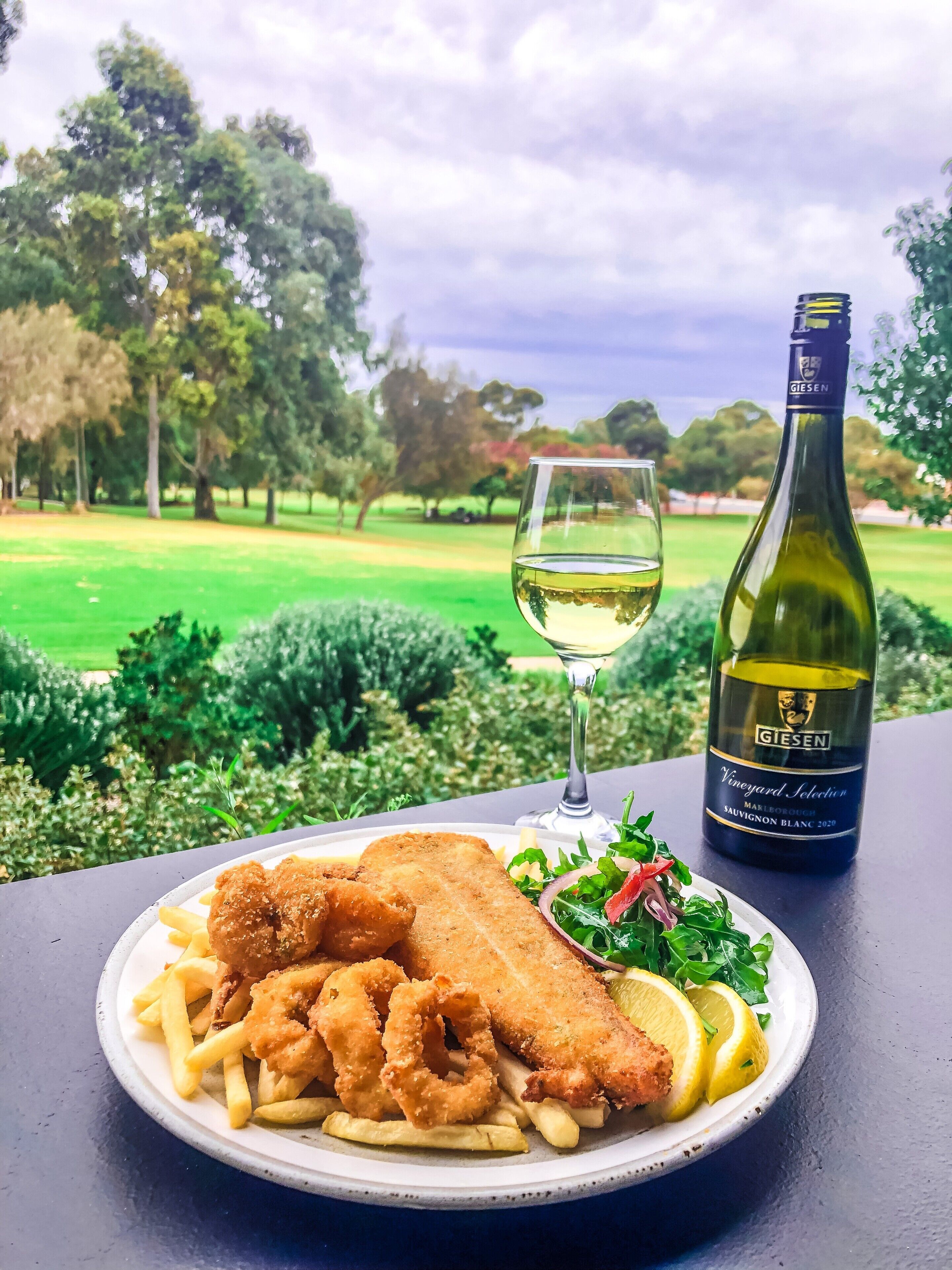 Un plato de pescado frito, papas fritas y aros de cebolla, junto a una botella de vino y una copa, sobre una mesa con vistas a un parque.