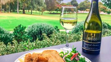 Un plato de pescado frito, papas fritas y aros de cebolla, junto a una botella de vino y una copa, sobre una mesa con vistas a un parque.