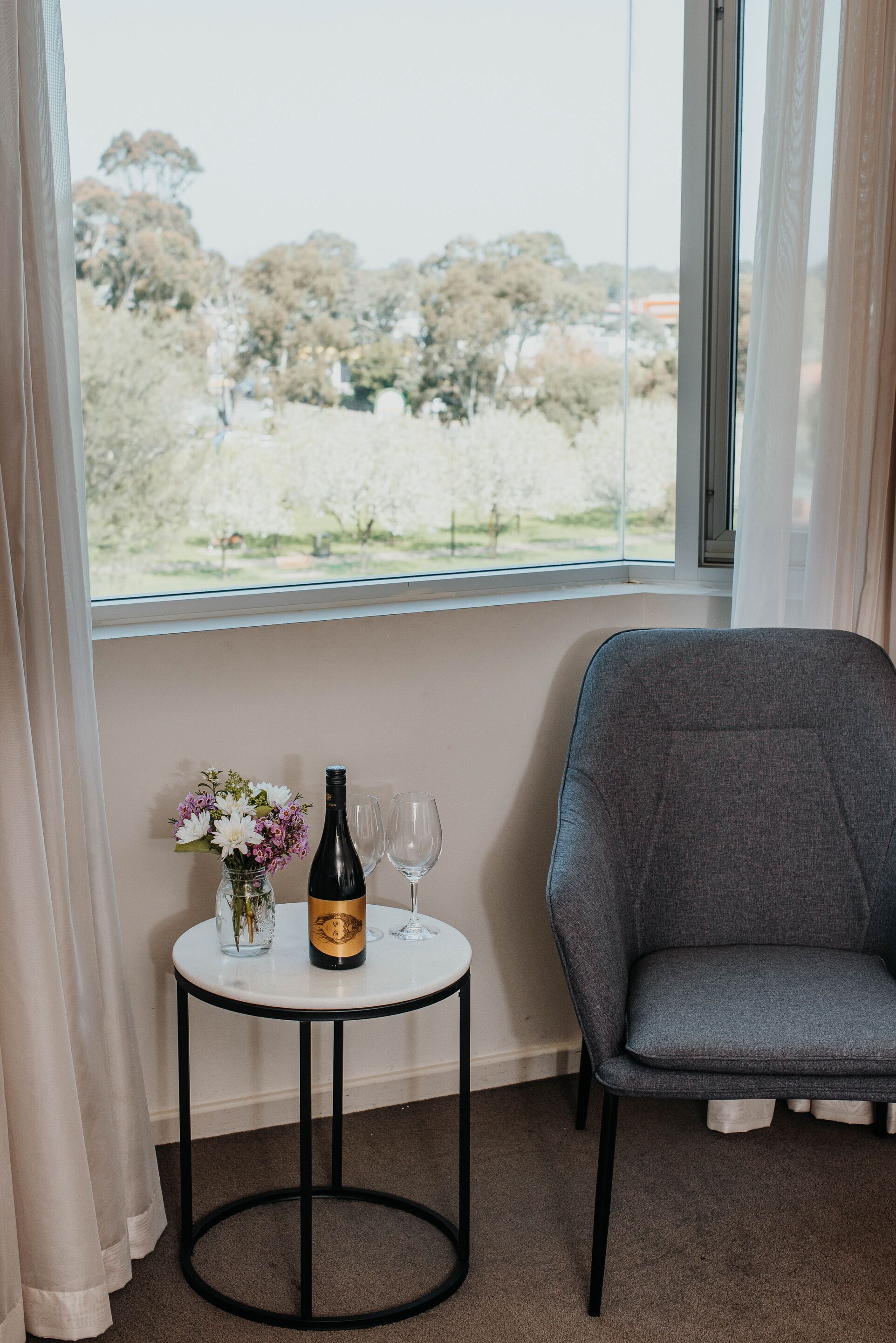A room with a large window, a small round table with a bottle and glasses, and a grey chair.