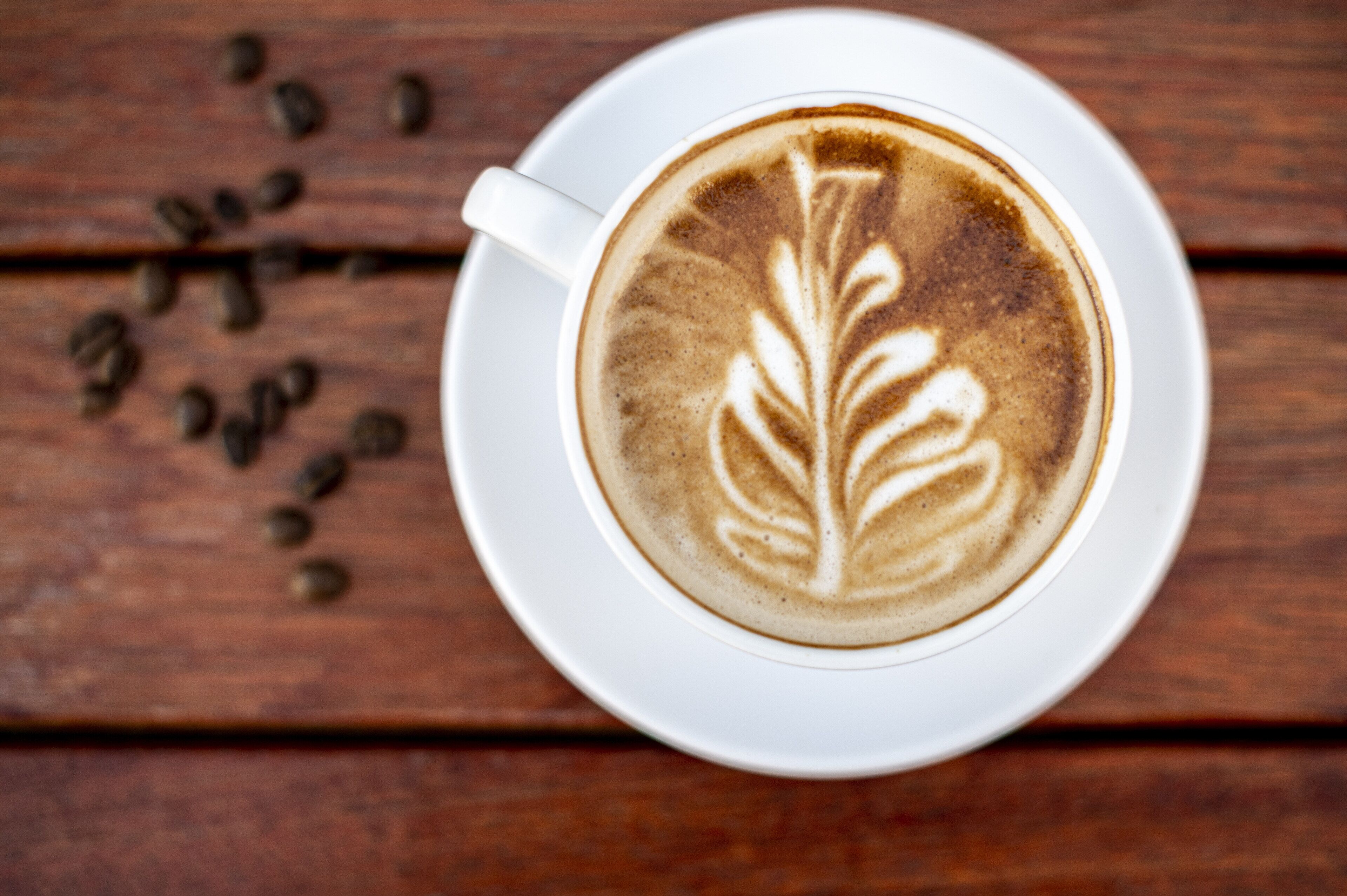 A cup of coffee with latte art on a white saucer, placed on a wooden surface with scattered coffee beans.