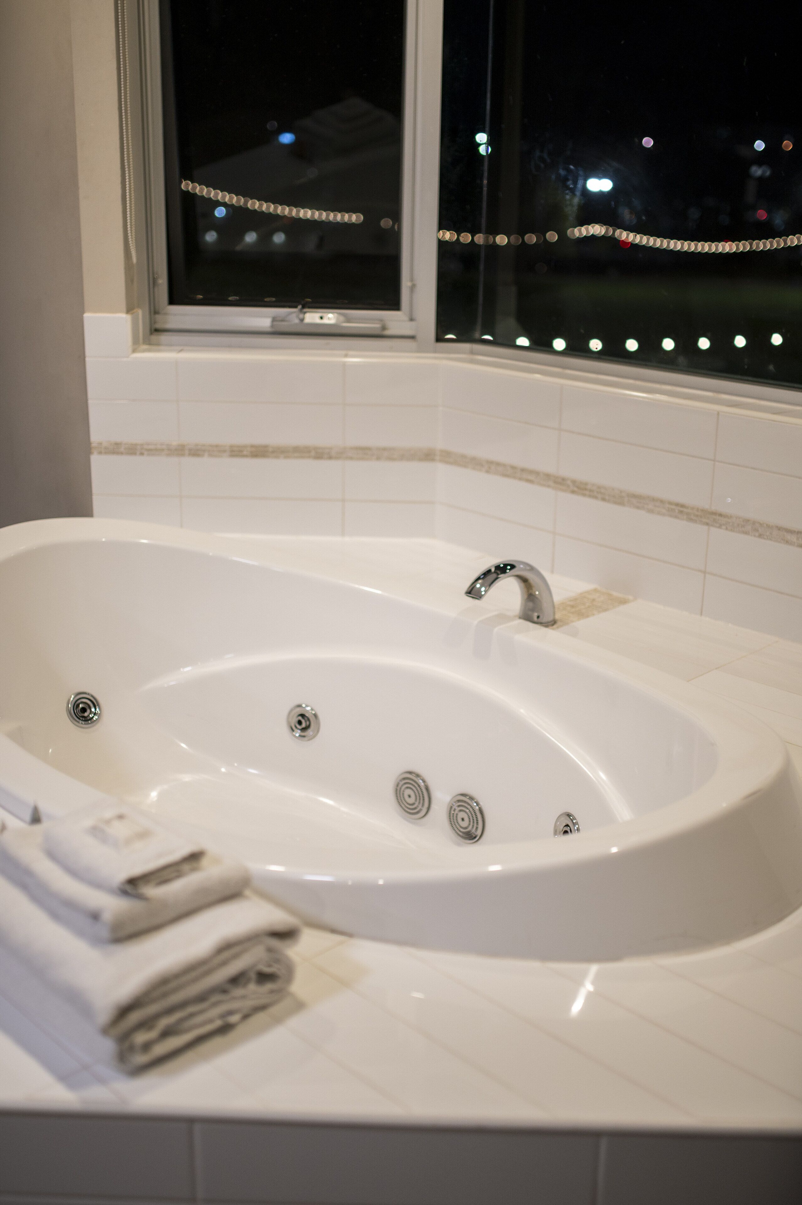 A modern bathroom with a large freestanding bathtub, a window with a view of the city at night, and a neatly arranged stack of towels.