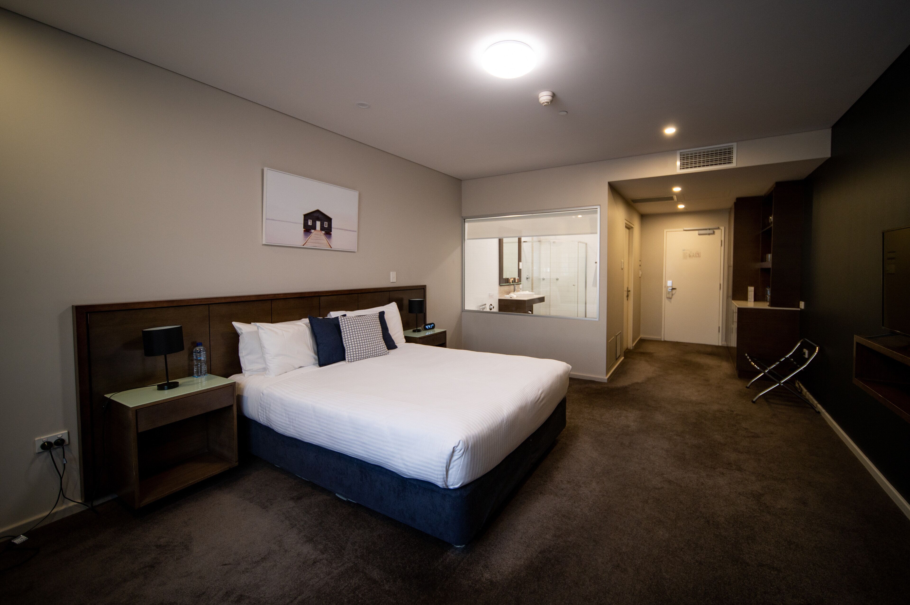 A hotel room with a large bed, a nightstand, a lamp, a water bottle, a mirror, a wardrobe, and a door leading to a bathroom.