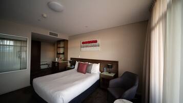 Una habitación de hotel con una cama grande, un escritorio, una silla, una mesita, una ventana con cortinas, un cuadro en la pared y una lámpara en el techo.