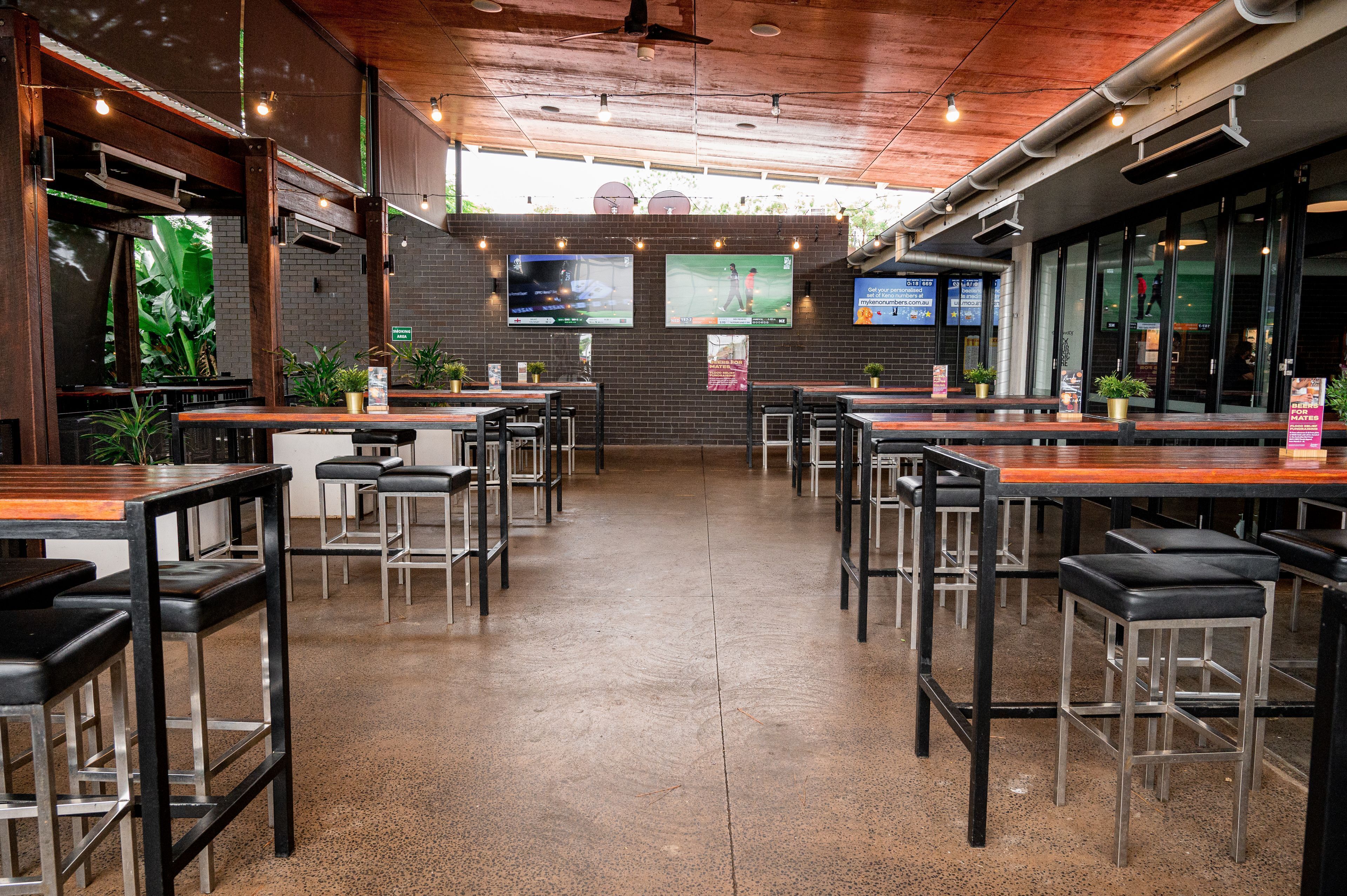 A restaurant with multiple tables and chairs, a brick wall, and two large screens displaying sports.
