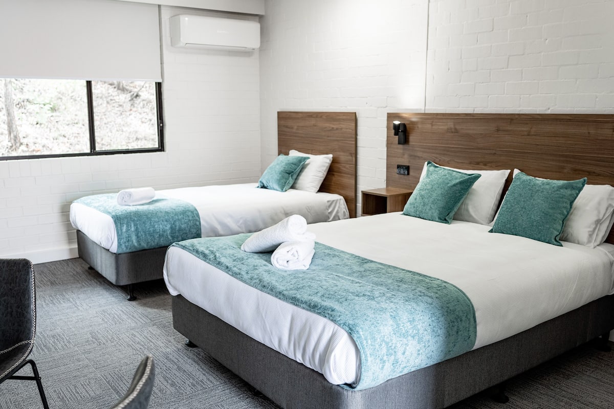 Habitación de hotel con dos camas, cada una con cabecero de madera, ropa de cama blanca y almohadones color turquesa. Incluye una mesita de noche con lámpara, una ventana con persianas y una silla.
