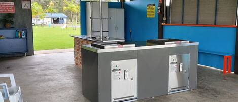 A covered outdoor area with a concrete floor, metal-framed ceiling, and a central counter with cabinets. There are signs indicating caution and surveillance.