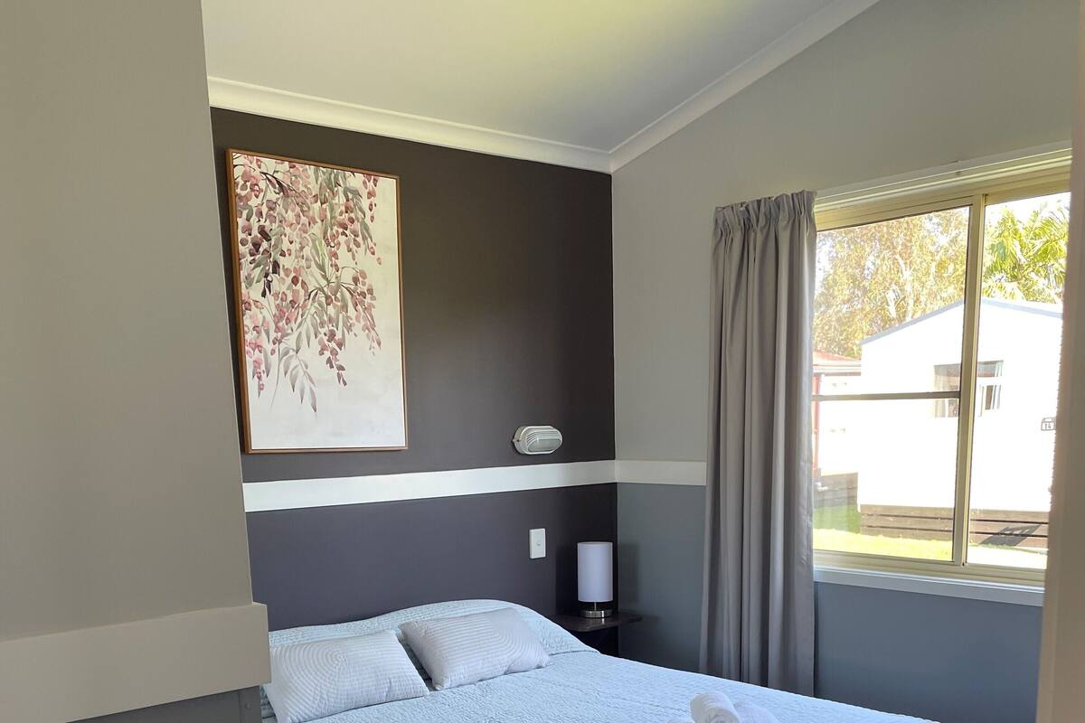A bedroom with a bed, a nightstand, a lamp, a painting, and a window with curtains.