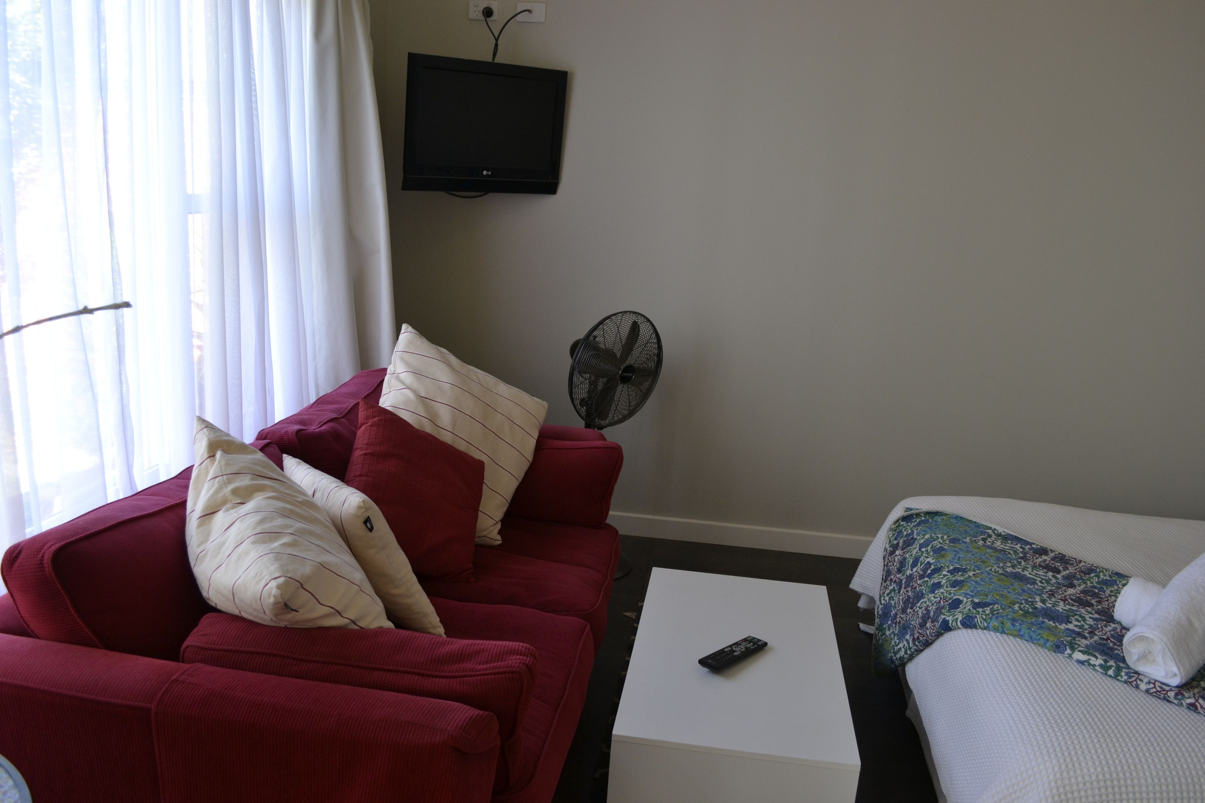 A room with a red sofa, a white coffee table, a bed with a patterned blanket, a TV, and a standing fan.