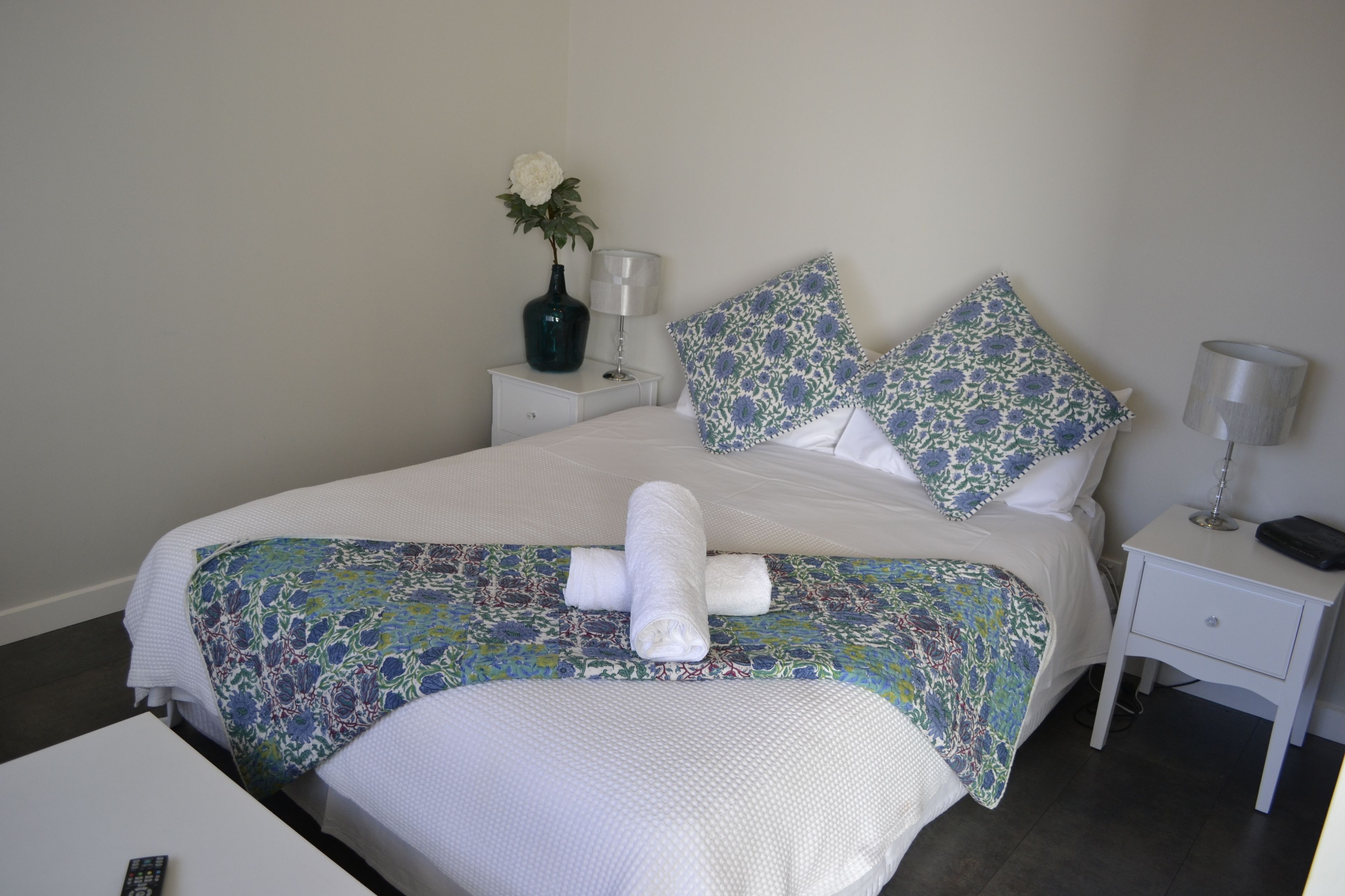 A neatly made bed with a floral bedspread, white pillows, and a vase with a flower on the nightstand.