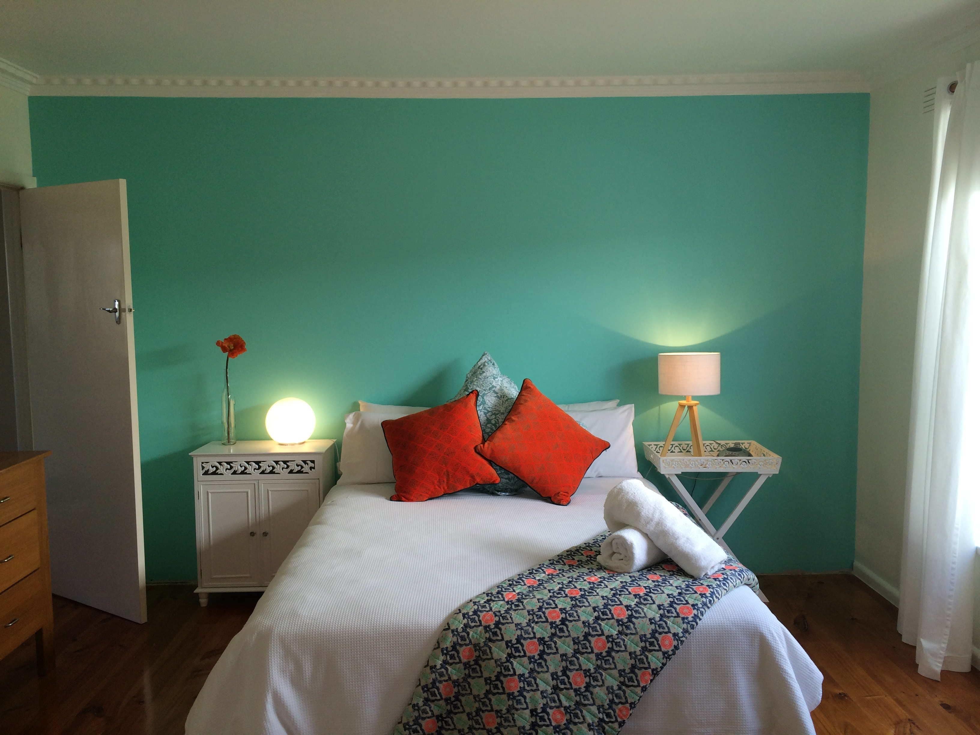 A bedroom with a teal wall, a white bed, red and grey pillows, a bedside lamp, and a nightstand with a vase and a lamp.