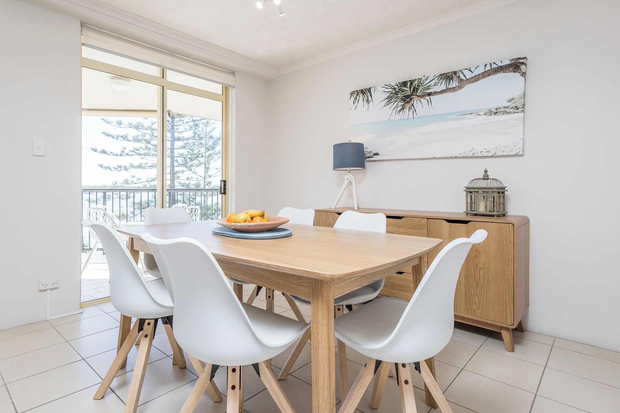 Un coin repas avec une table en bois et des chaises blanches, un saladier rempli de fruits, et une vue sur l’extérieur grâce à de grandes fenêtres.