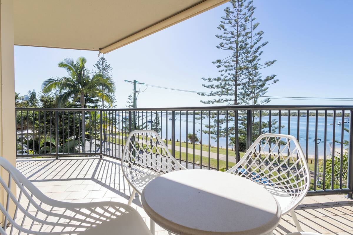 A balcony with two white chairs and a small table, overlooking a beach and palm trees.