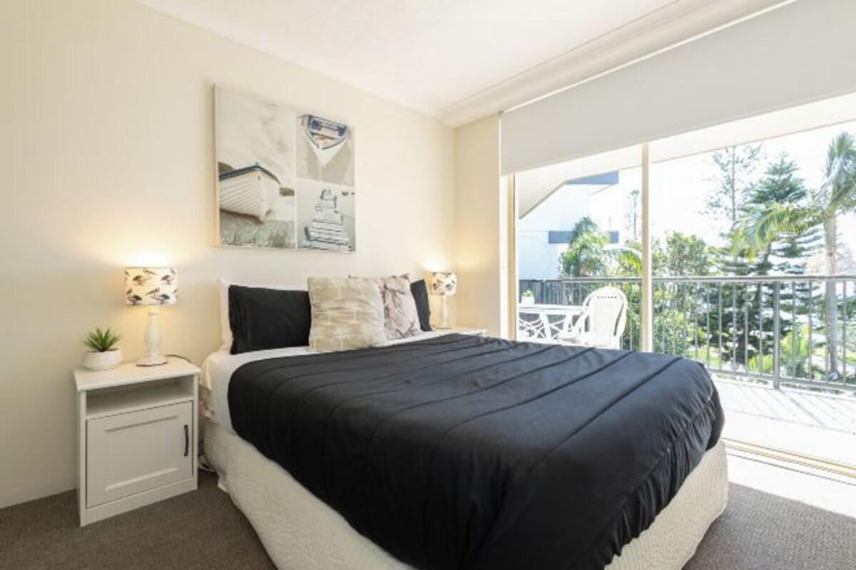 Un dormitorio con una cama grande, una mesita de noche con lámpara, un cuadro en la pared y un balcón con vistas a palmeras.
