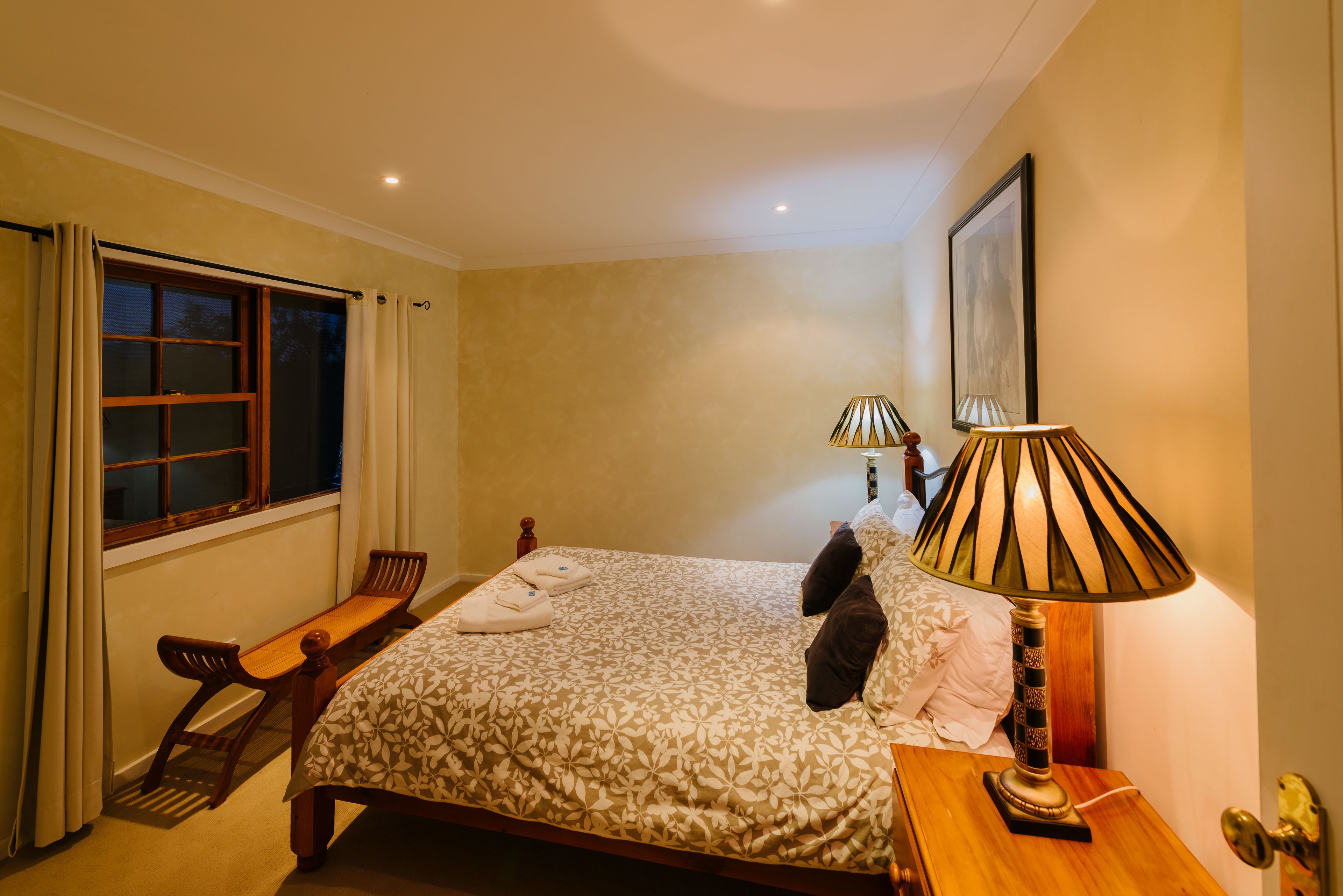 A bedroom with a bed, bedside lamps, a window with curtains, a wooden bench, and a door.