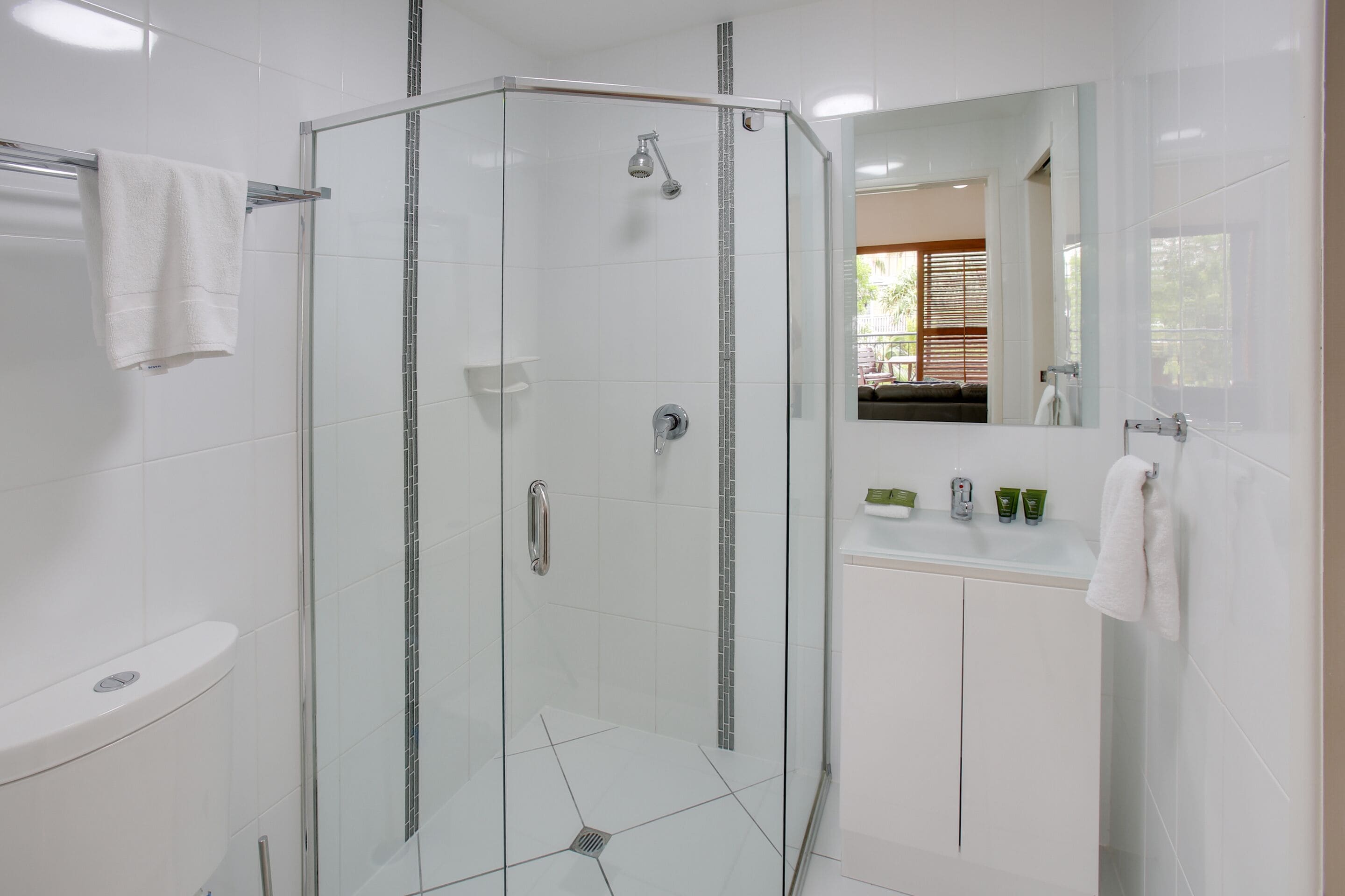 Un baño moderno con ducha envuelta en vidrio, azulejos blancos, tocador con artículos de aseo y un espejo grande.
