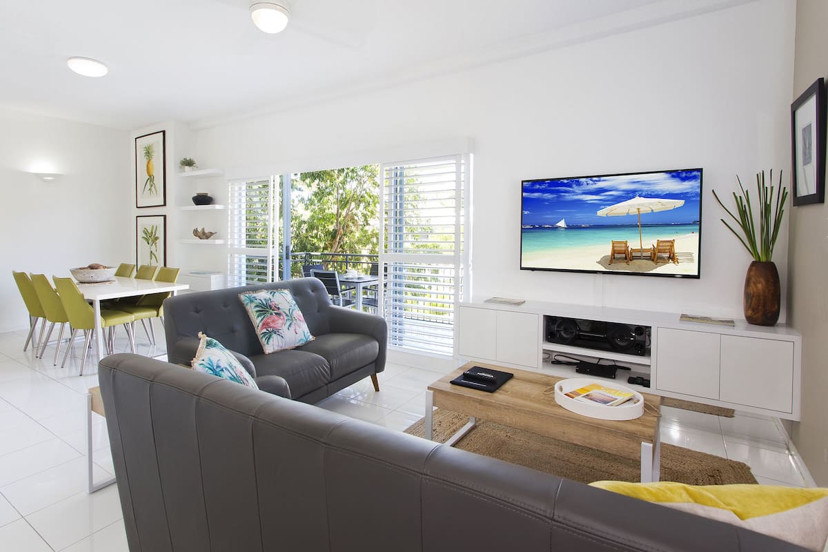 A modern living room with a grey sofa, dining area with green chairs, a TV on the wall, and a view of the outdoors through large windows.