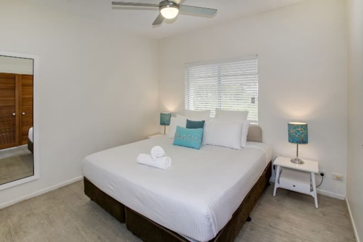 A bedroom with a bed, a ceiling fan, a window with blinds, a nightstand with a lamp, and a mirror on the wall.