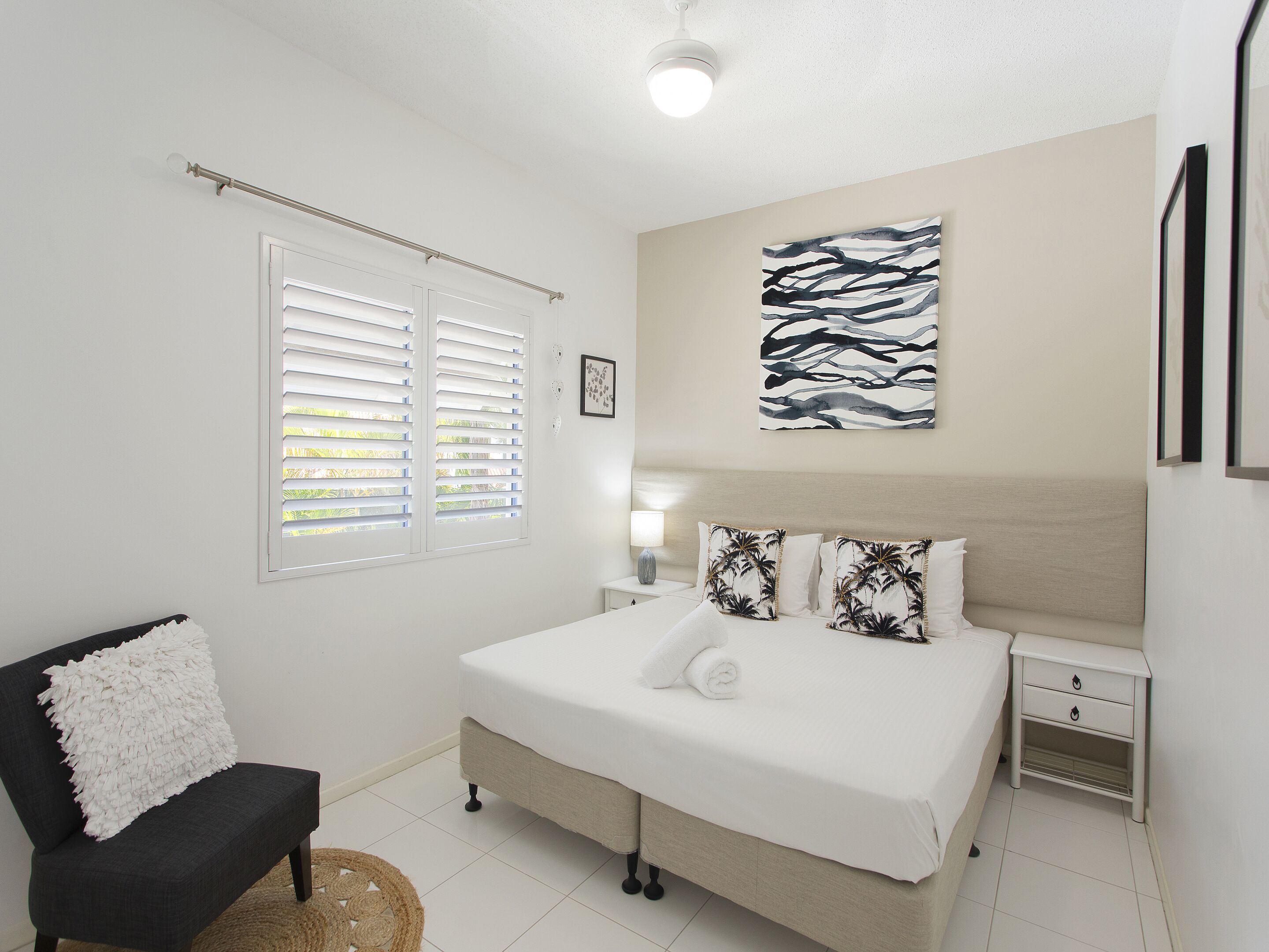Quarto com cama, cadeira, mesa de cabeceira, luminária e janela com persianas.