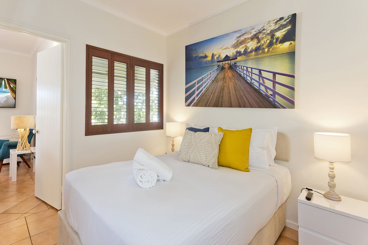 A hotel room with a large bed, a painting of a pier, a wooden door, and a nightstand with a lamp.