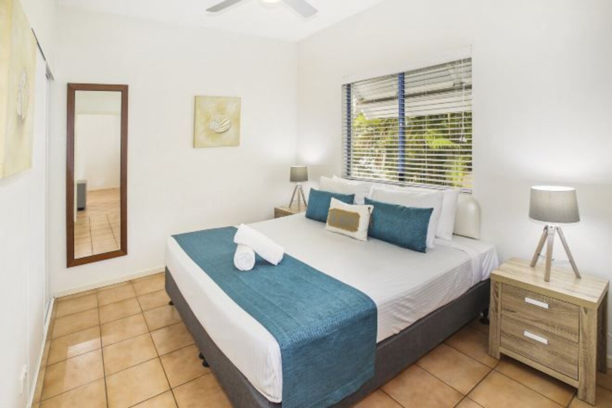 A bedroom with a bed, bedside tables, a window with blinds, a mirror, and a ceiling fan.