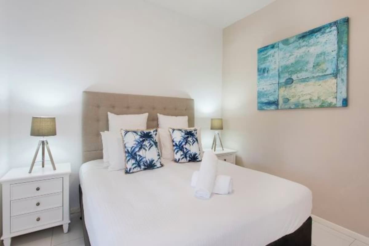 A neatly made bed with white bedding and two decorative pillows, a bedside table with a lamp, and a wall-mounted abstract painting.