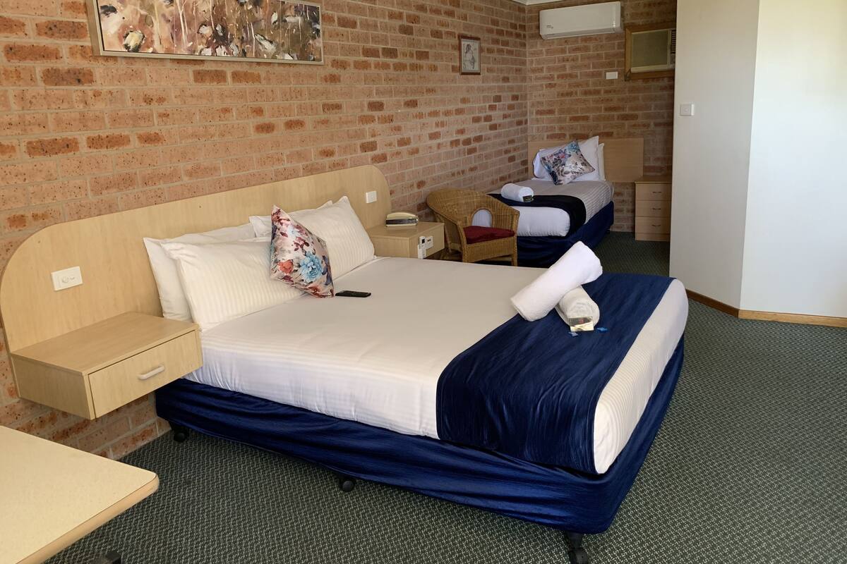 A hotel room with a brick wall, a large bed with a blue bedspread, a wooden nightstand, a wicker chair, and a small table.