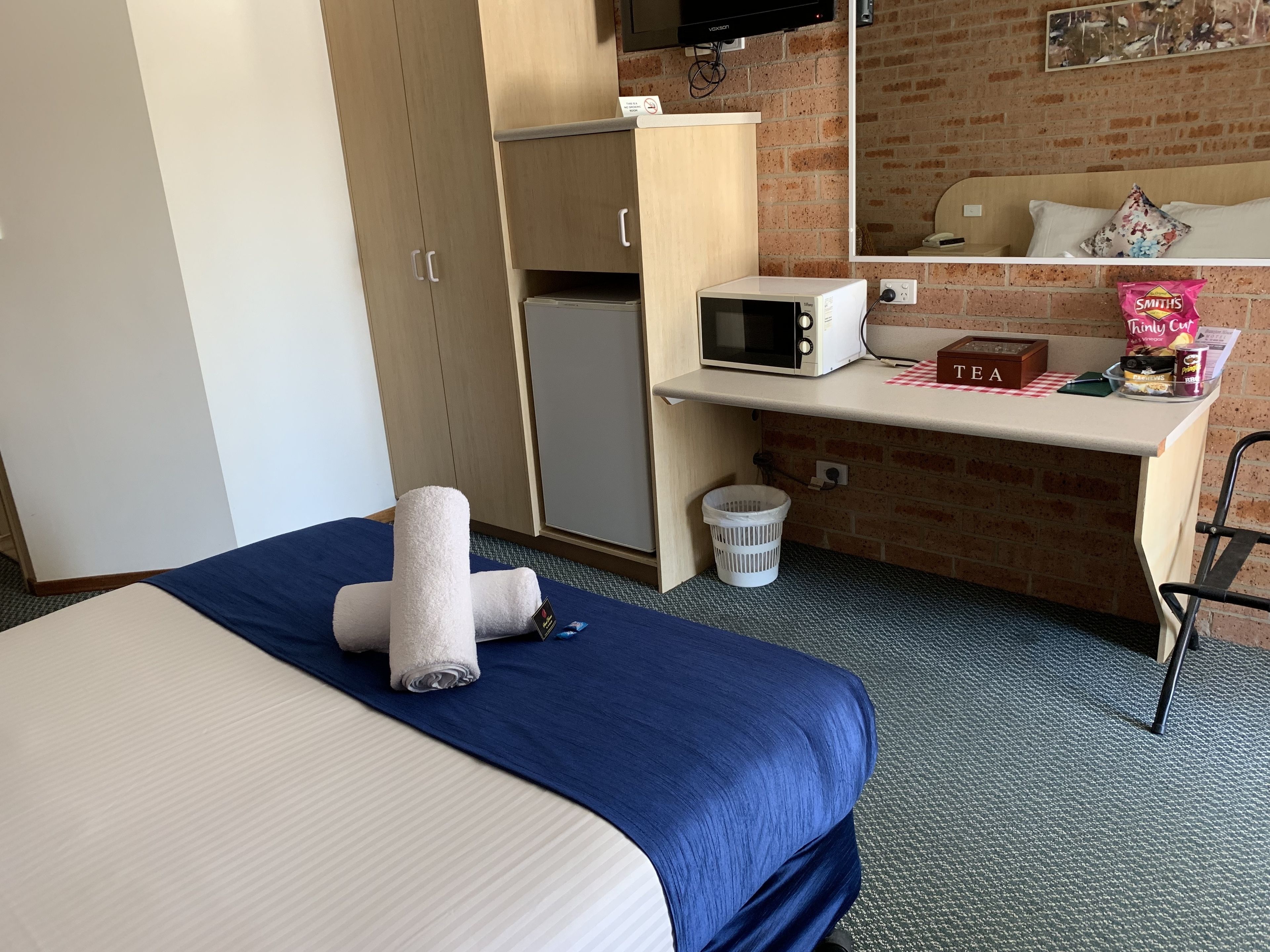 A hotel room with a bed, a desk with a microwave, a tea station, and a trash bin.