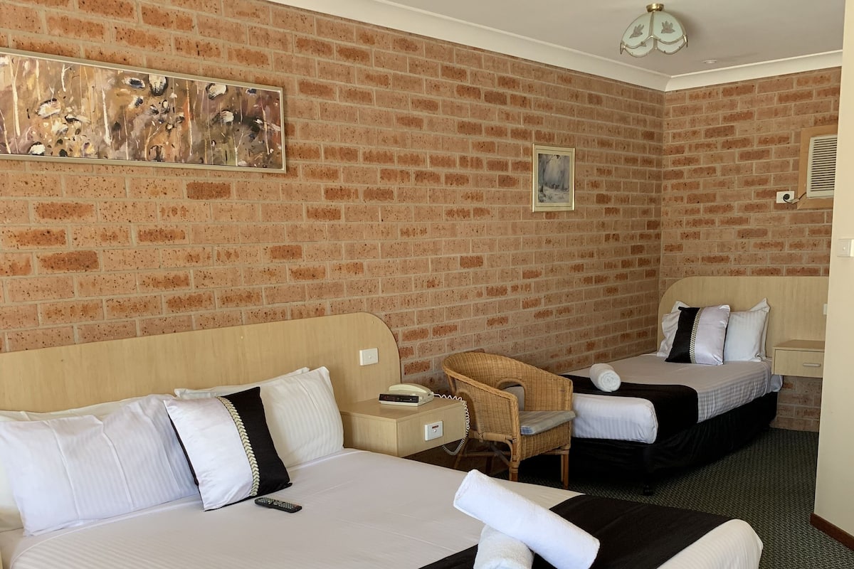 A hotel room with two beds, a brick wall, a ceiling fan, and a small table with a chair.