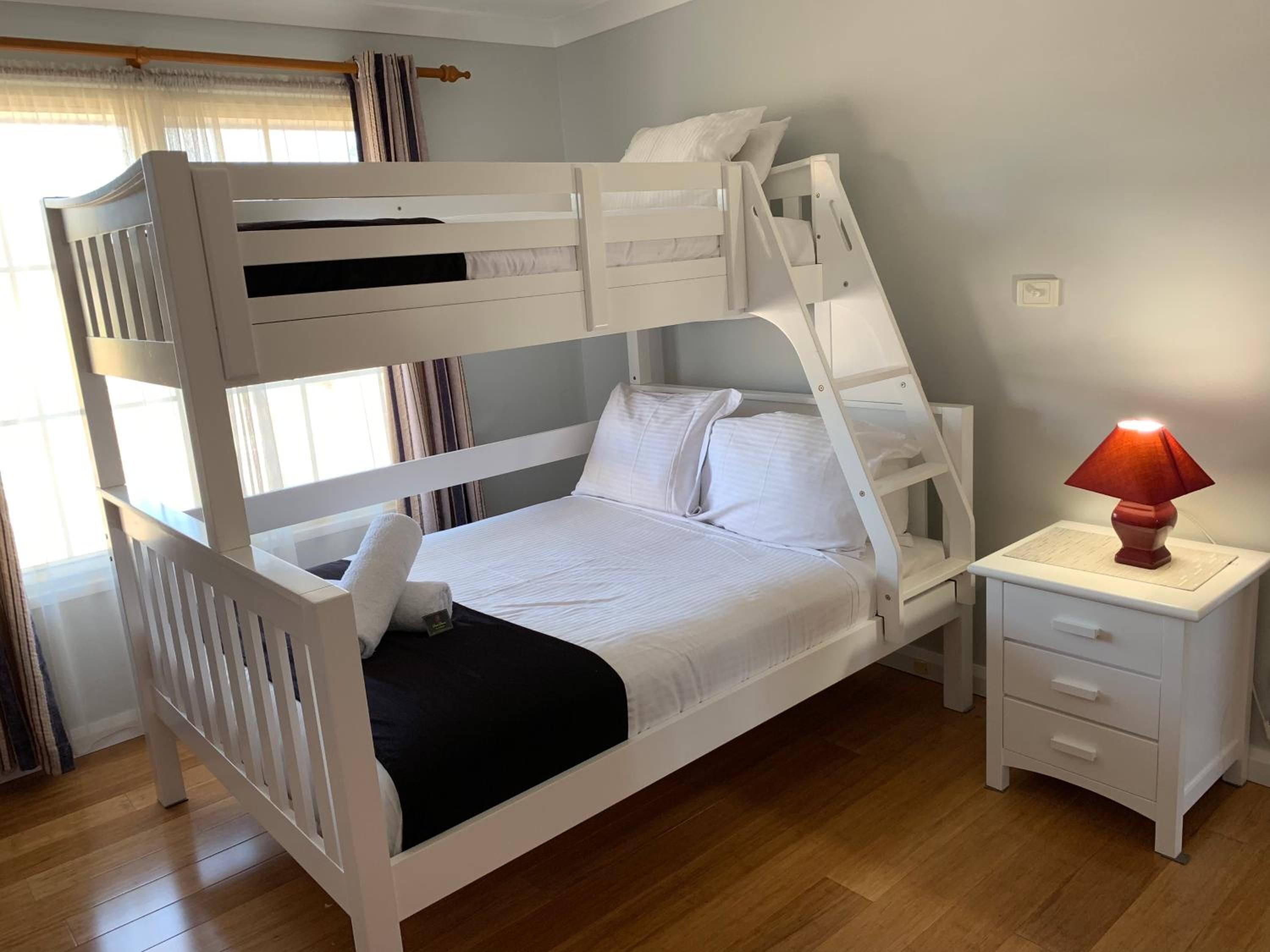 A bunk bed with a white frame, a bedside table with a lamp, and a window with curtains.