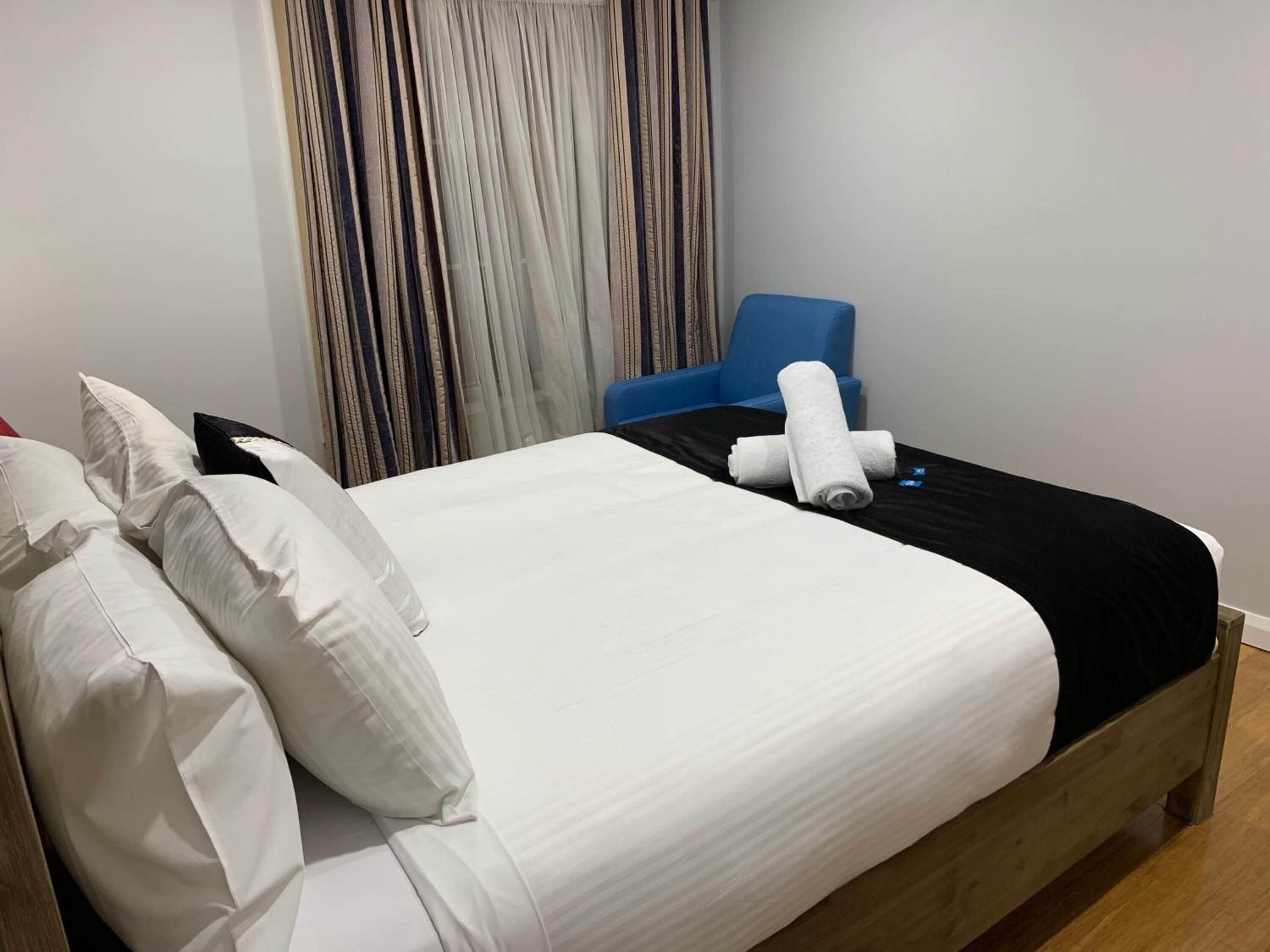 A neatly made bed with white linens and a black bedspread, a blue chair, and a curtain with a striped pattern.