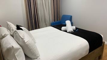 A neatly made bed with white linens and a black bedspread, a blue chair, and a curtain with a striped pattern.