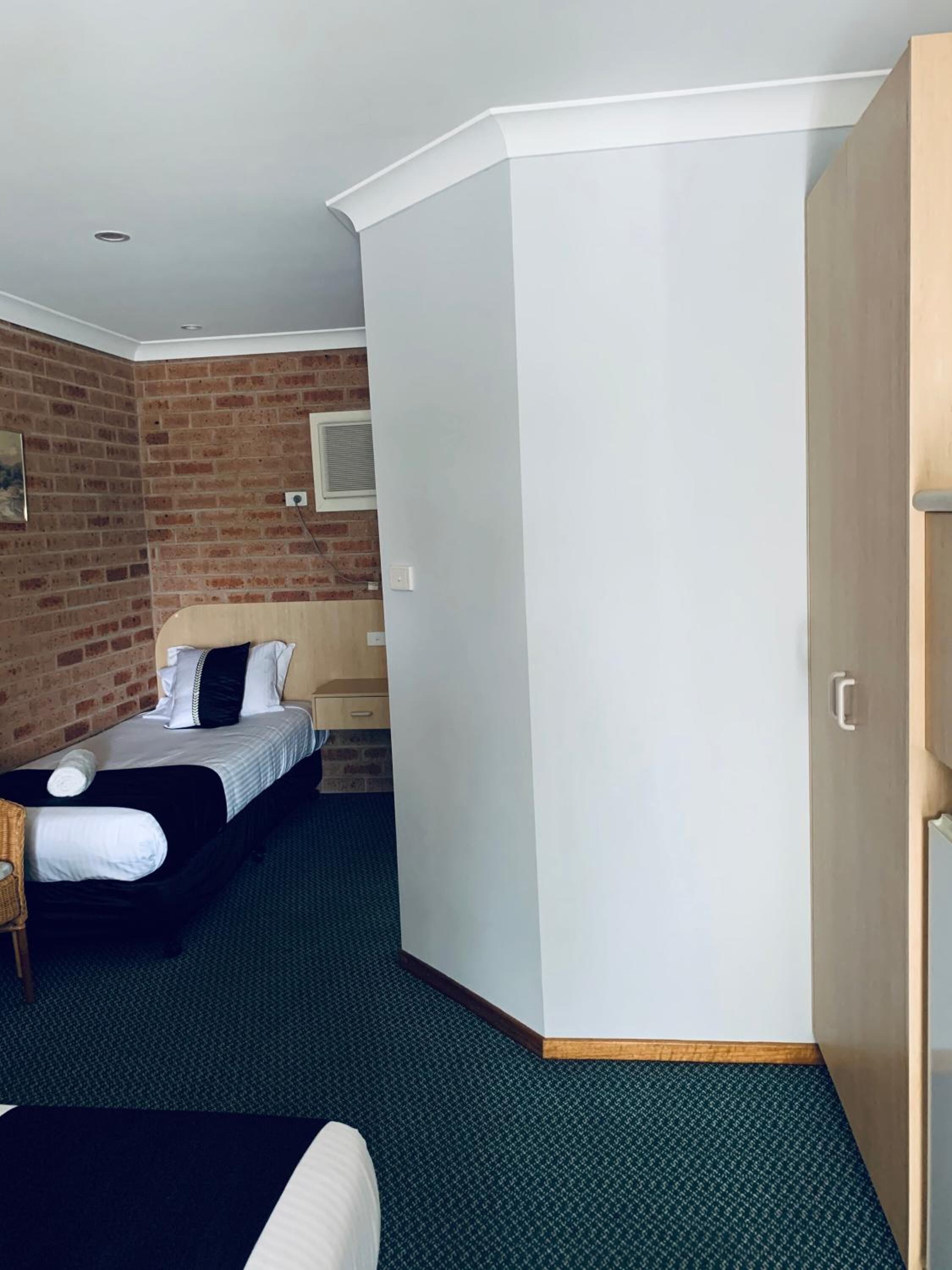 A hotel room with a brick wall, a bed with white bedding, a wooden wardrobe, and a green carpet.