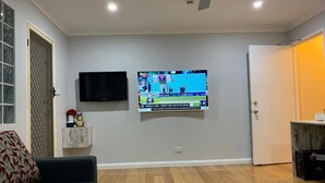 A living room with a flat-screen TV mounted on the wall, a grey sofa, a coffee table with a plant, and a wooden floor. - Branxton House Motel, Hunter Valley (Branxton)