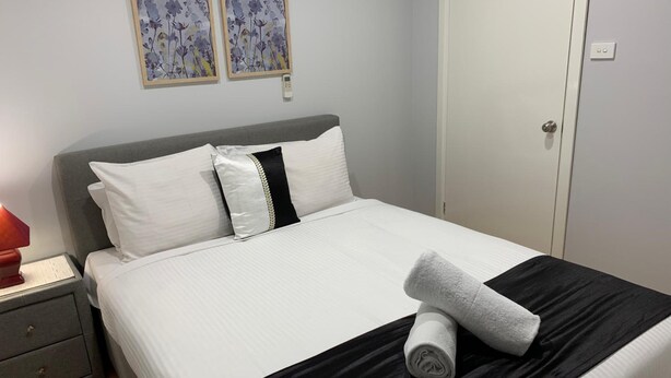 A neatly made bed with white bedding and a black accent, a grey nightstand with a red lamp, and two framed floral artworks on the wall. - Branxton House Motel, Hunter Valley (Branxton)