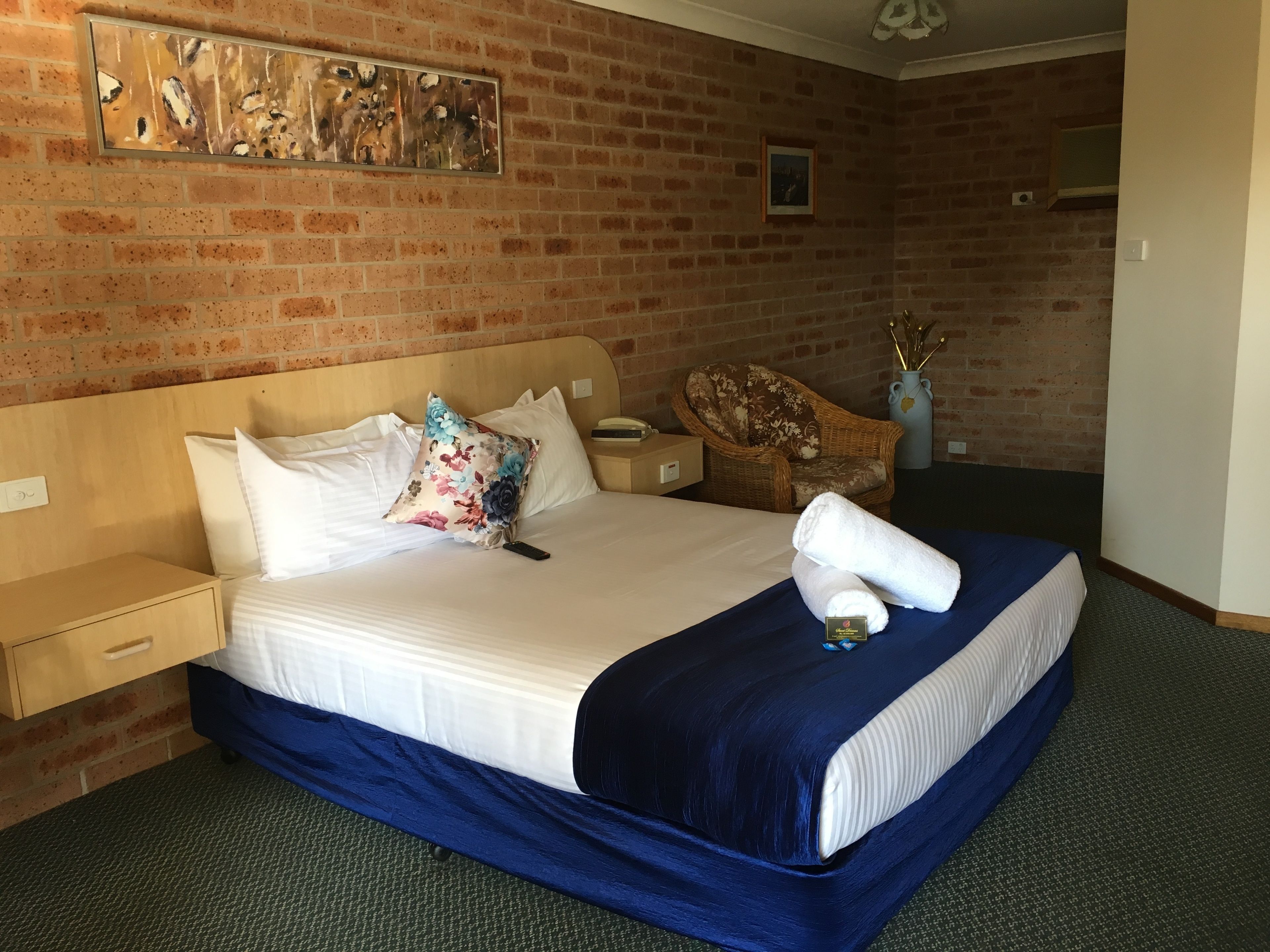 Una habitación de hotel con pared de ladrillo, cama con ropa blanca y colcha azul, mesita de noche de madera, silla de mimbre y un cuadro en la pared.
