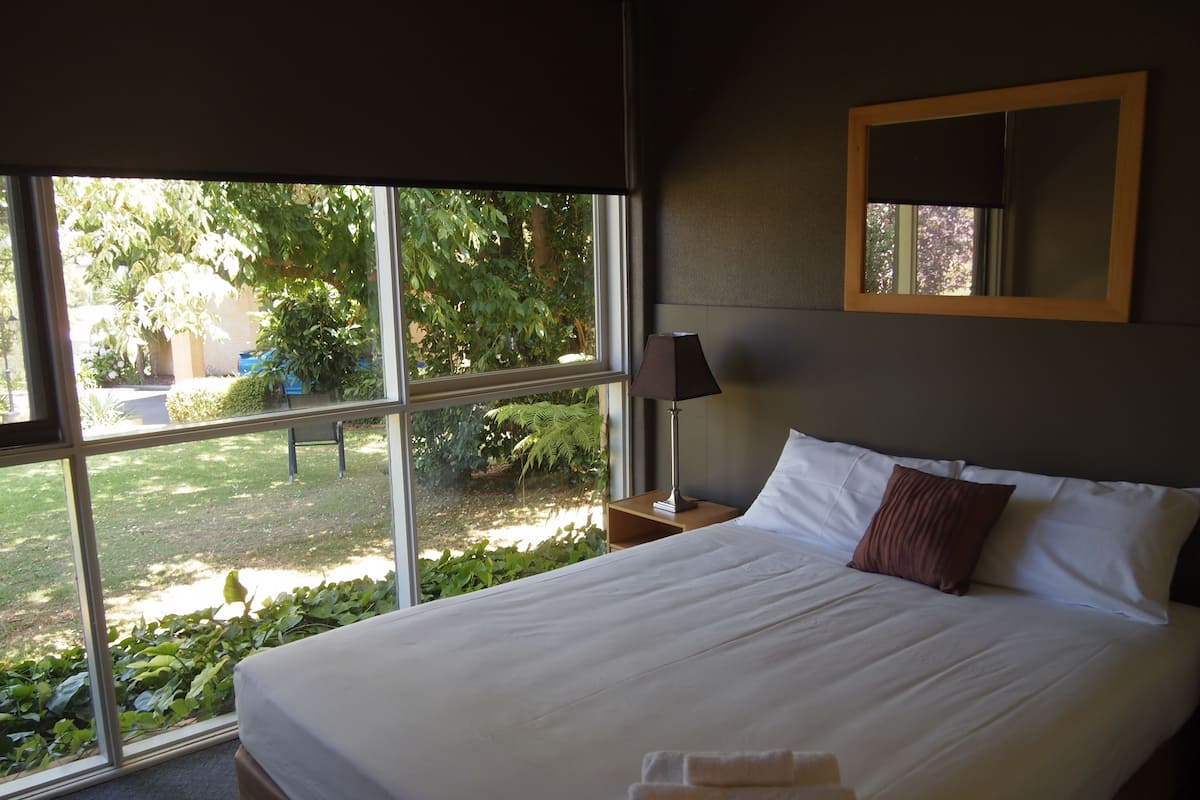A neatly made bed with white linens and a brown pillow, a bedside lamp, and a window with a view of a garden.