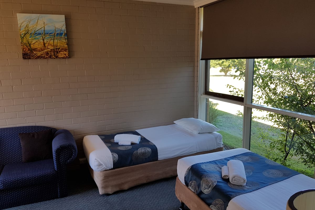 A hotel room with two beds, a large window with a view of greenery, a brick wall, and a painting.
