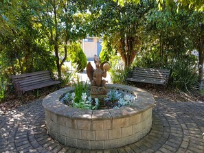 A circular stone fountain with a statue of a figure holding a mushroom, surrounded by a brick border. Two wooden benches are placed on either side of the fountain.