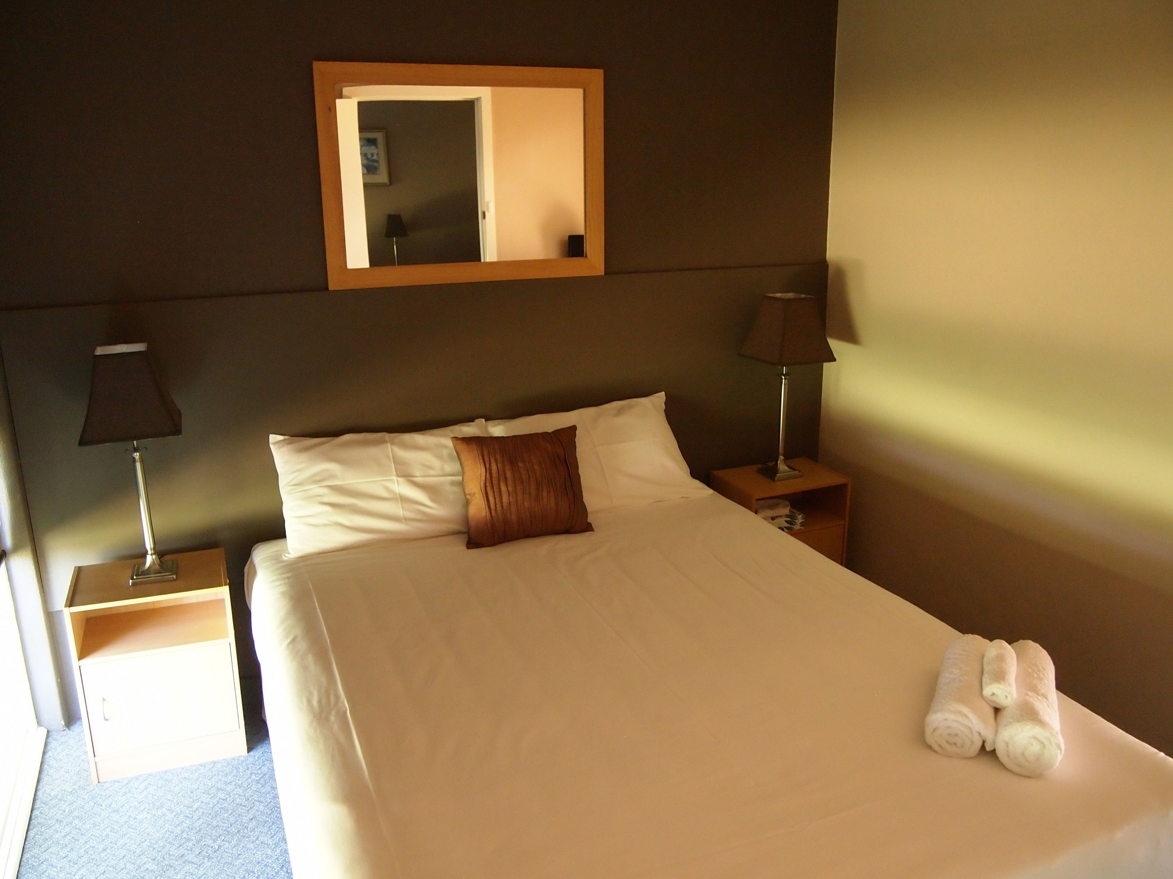 A neatly made bed with white linens, a brown pillow, and two rolled towels on the side. There are two bedside tables with lamps, one on each side of the bed.
