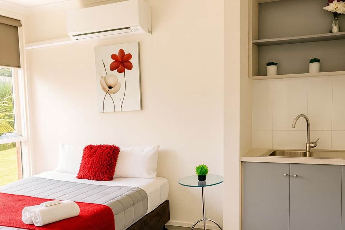 A bedroom with a bed, a red throw, a small table with a plant, a wall-mounted air conditioner, and a kitchen area with a sink and cabinets.