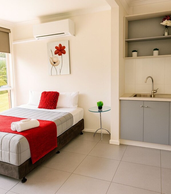 A bedroom with a bed, a red throw, a small table with a plant, a wall-mounted air conditioner, and a kitchen area with a sink and cabinets.