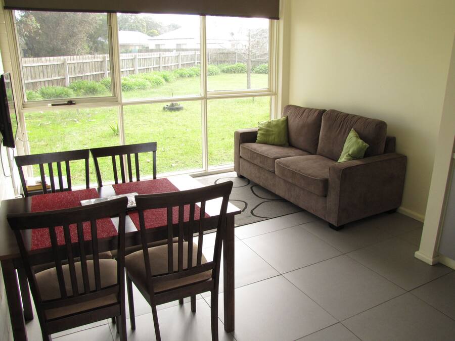 A living room with a brown sofa, green cushions, a dining table with chairs, and a view of a backyard through large windows.